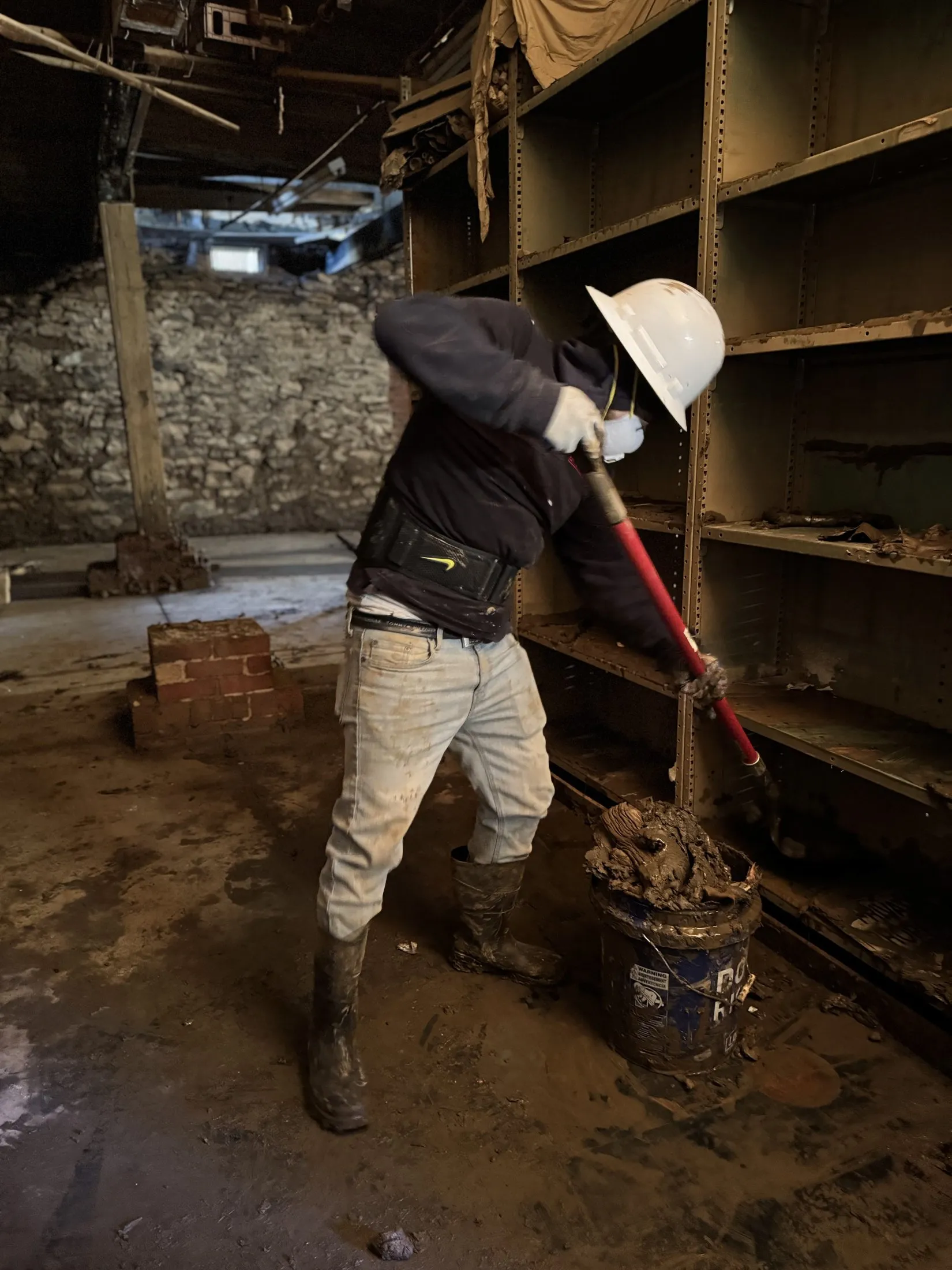 A Carolina Pro Restoration technician in a hard hat and respirator shoveling flood mud out of a commercial building